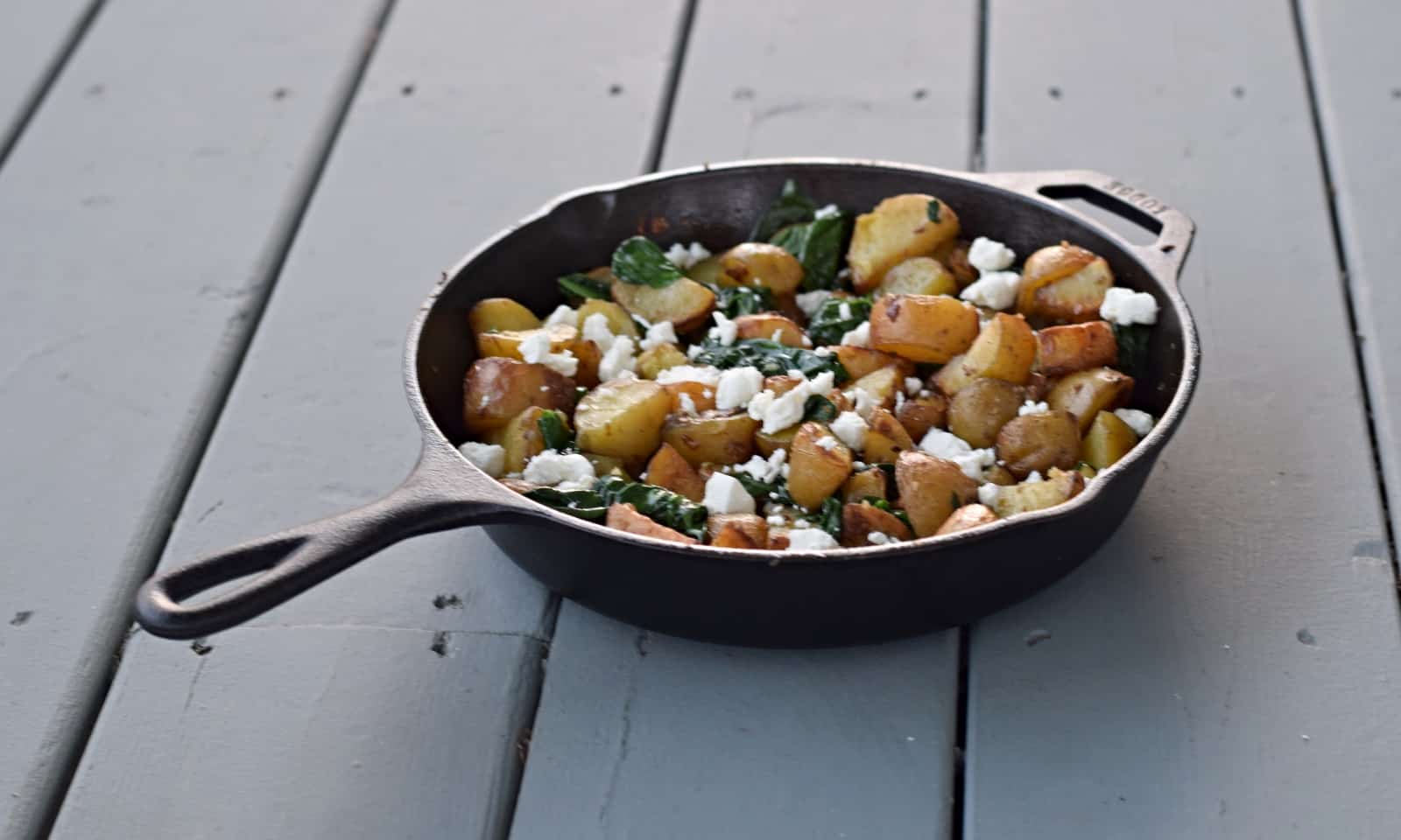 This photo shows a cast iron skillet with oven roasted potatoes with spinach and garlic on a blue wooden background.