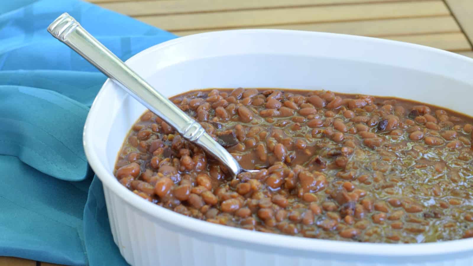 Image shows a white casserole dish filled with homemade baked beans on wooden table with a silver spoon sticking out from it.