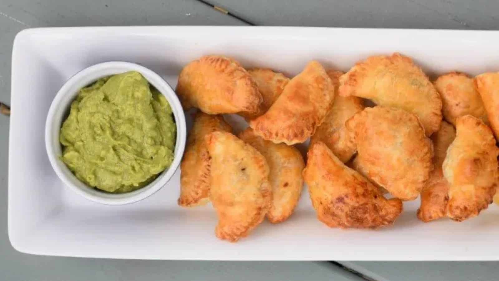 Image shows an overhead shot of Puff Pastry Beef Empanadas on a white tray with a bowl of guacamole to the left of them.