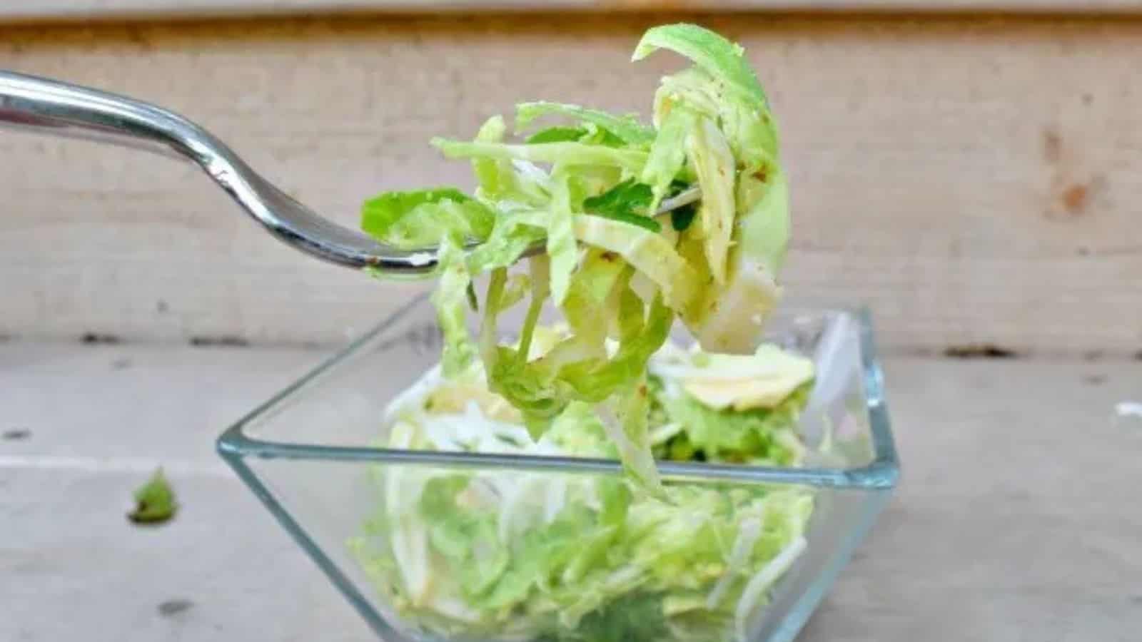 Image shows a fork holding a bite of shaved brussels sprouts salad with a clear bowl containing the rest of the serving behind it.