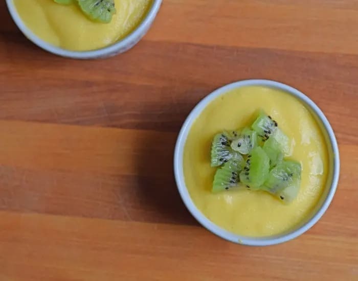An overhead shot shows two ramekins with mango custard topped with chopped kiwi on a wooden cutting board.