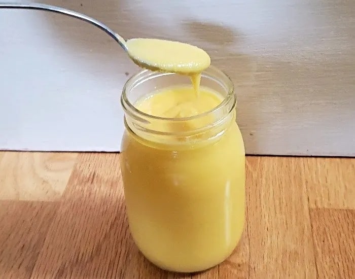 A front on photo shows mango curd in a glass jar with a spoon that has removed some curd.