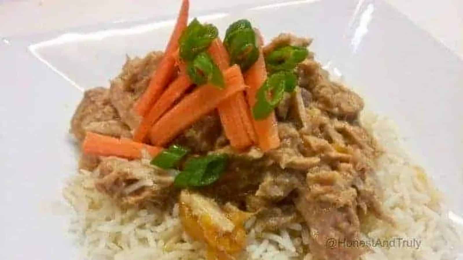 Image shows Slow Cooker Thai Pork on a bed of rice on a white plate with sliced carrots and green onion on top of it.