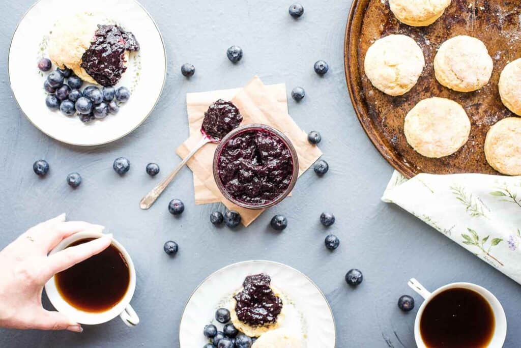 Small batch blueberry jam featured in the middle of the table with biscuits, coffee, and serving plates.