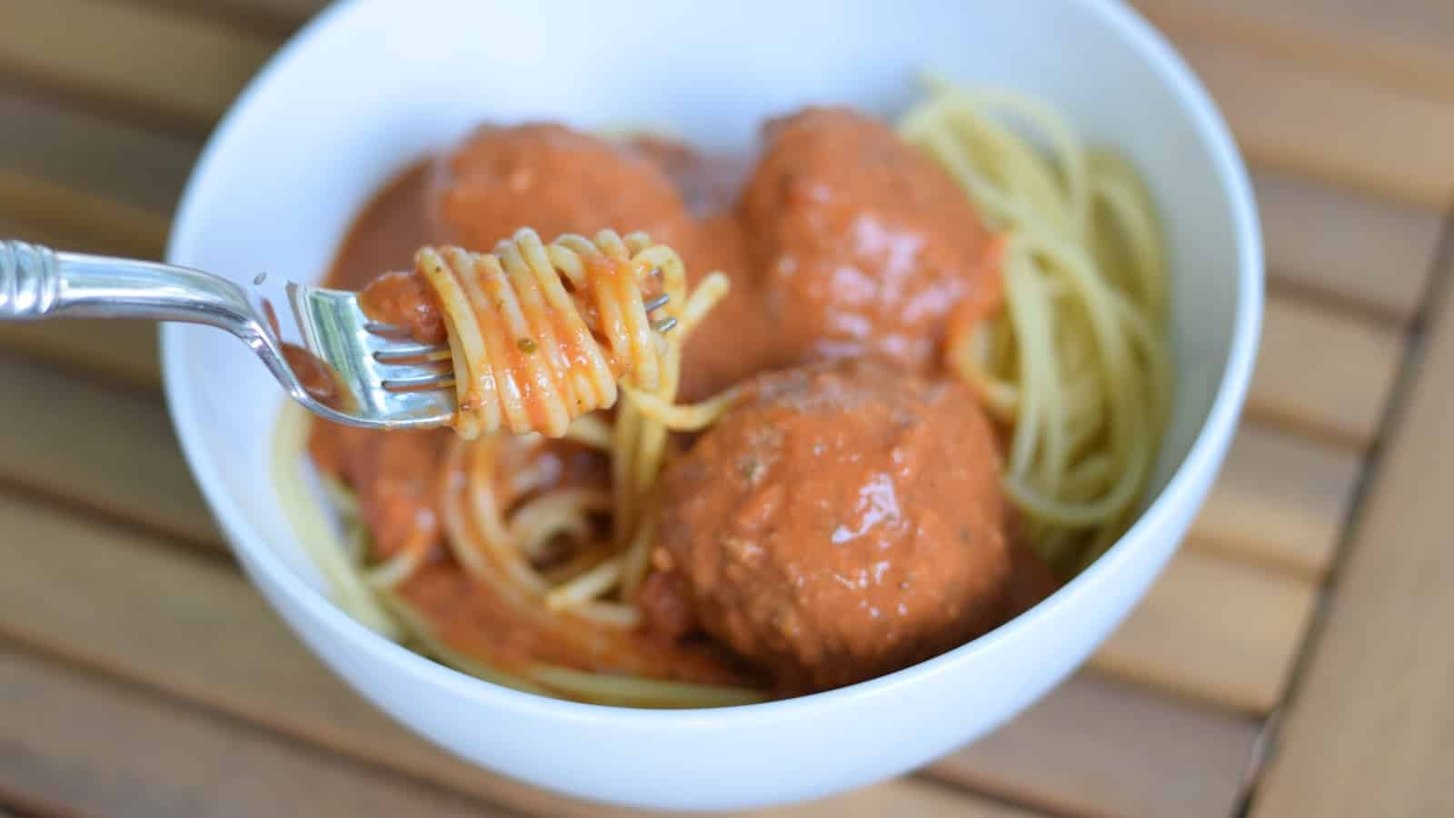 Image shows Spaghetti and meatballs with a fork twirling pasta in a white bowl.