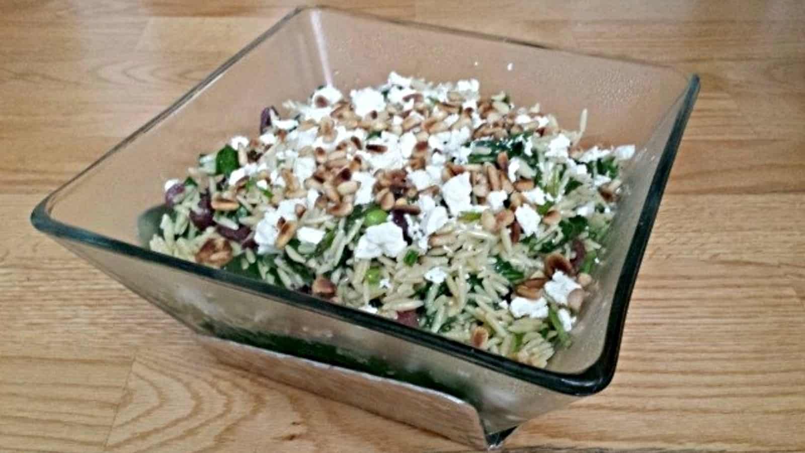 This photo shows a spinach orzo salad in a square glass bowl sitting on a black granite countertop.