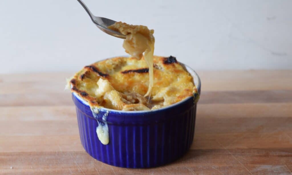Image shows a Spoon pulling cheese from a bowl of French onion soup in a blue ramekin.
