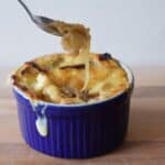 Image shows a Spoon pulling cheese from a bowl of French onion soup in a blue ramekin.