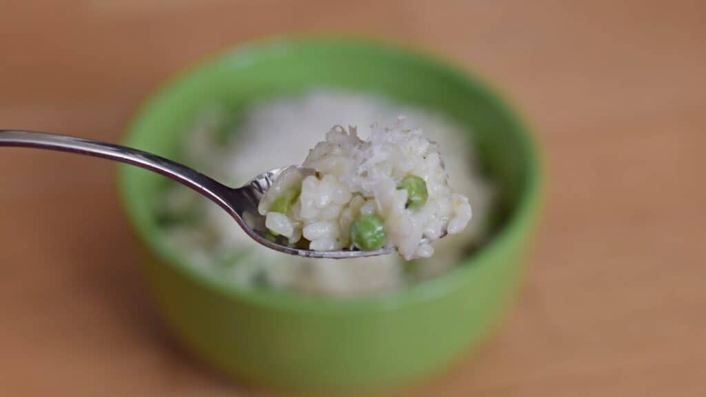 Image shows a Spoonful of risotto with peas and lemon with the rest of the bowl in the background.