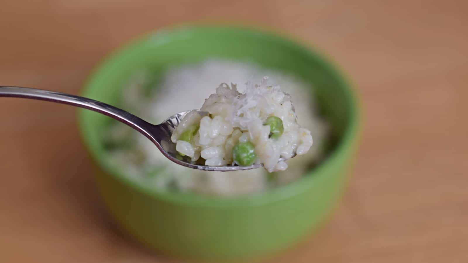 Image shows a Spoonful of risotto with peas and lemon with the rest of the bowl in the background.