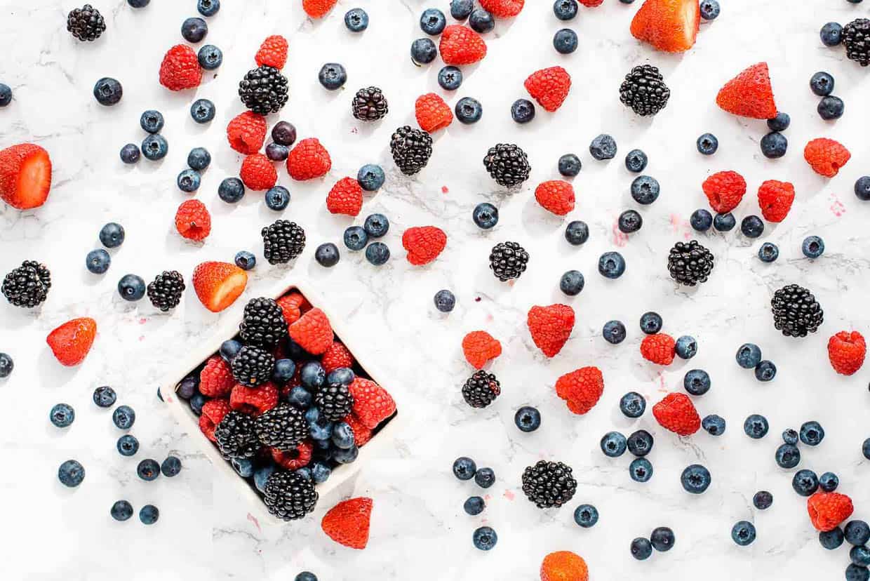Raspberries, blueberries & blackberries spread out on a marble surface with a carton of berries in the bottom left corner.