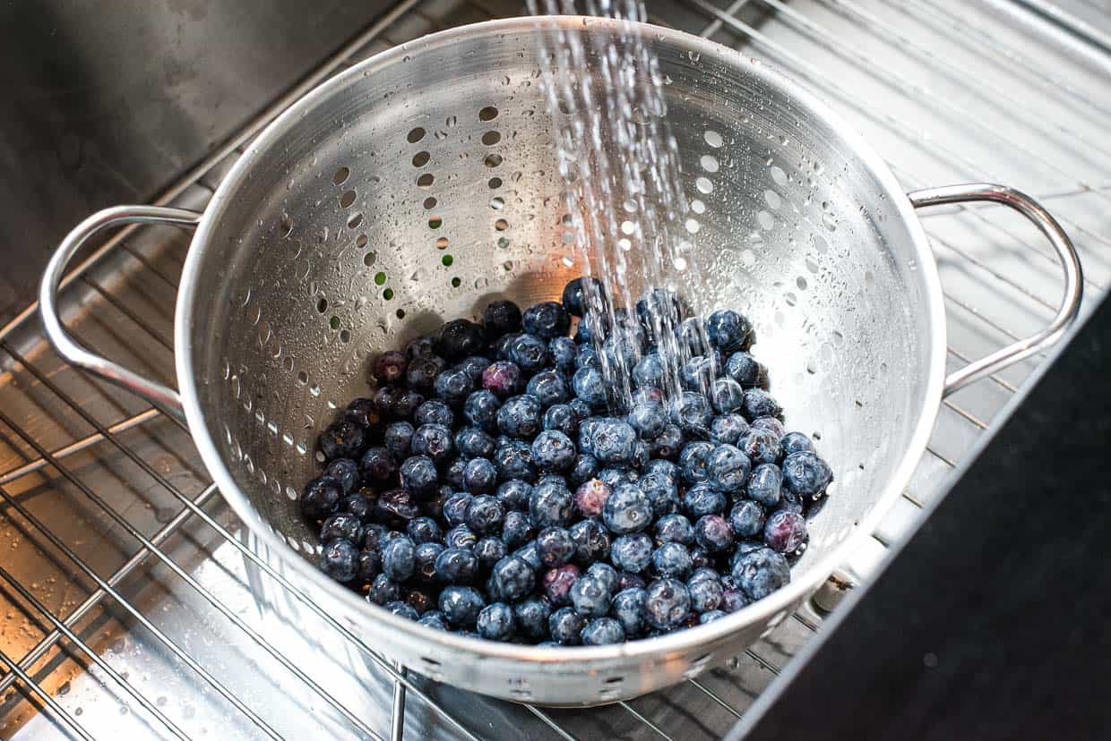 Blueberries in a silver colander being rinsed in a kitchen sink.