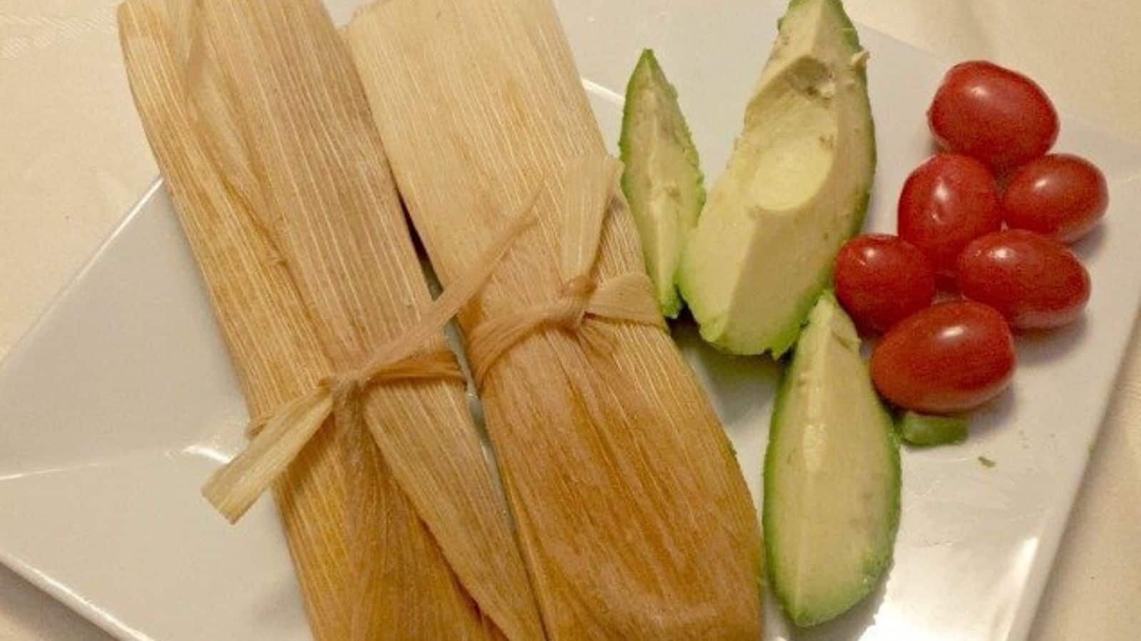Image shows two Tamales sitting on a white plate with slices of avocado and tomatoes next to it.