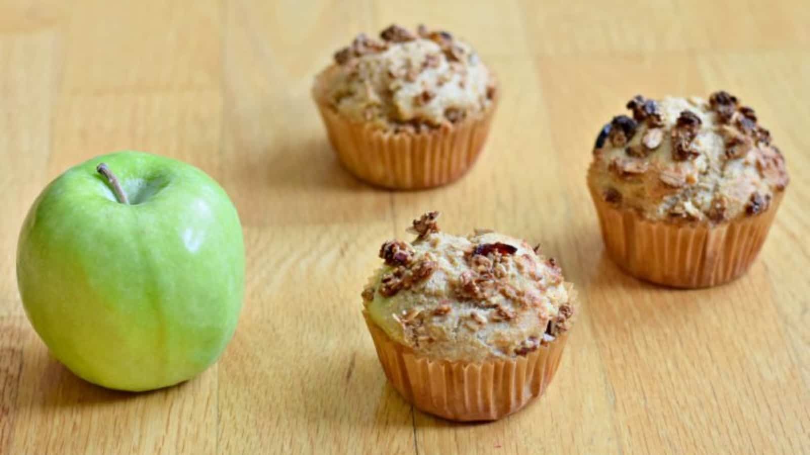 Three apple cinnamon muffins on a wooden table with an apple.