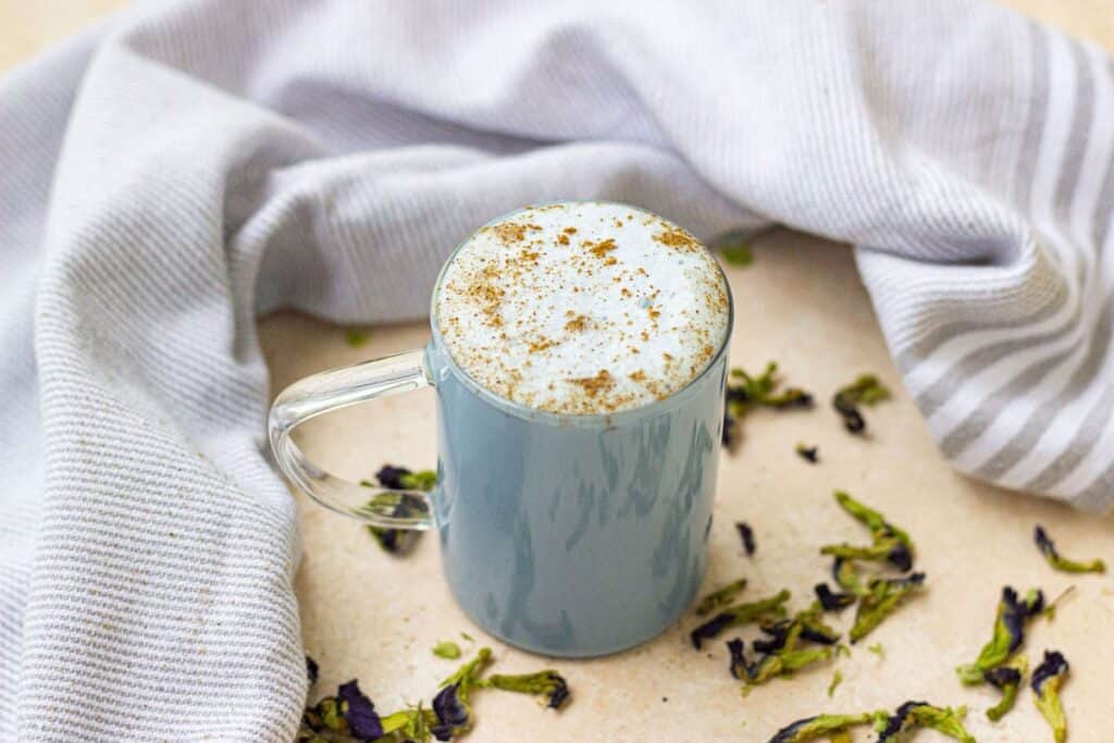 Blue matcha latte in a glass mug with a sprinkle of ground cinnamon as garnish. Dried butterfly pea flowers in background.