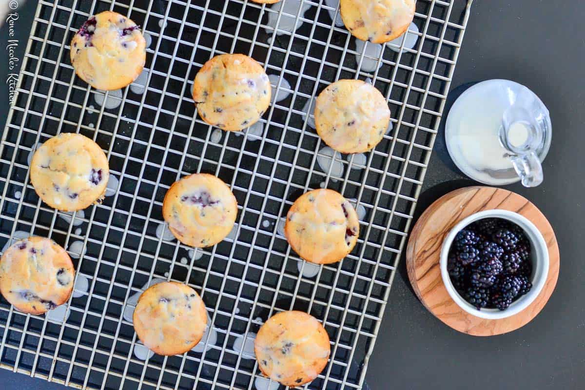 Blackberry muffins drizzled with lemon glaze sitting on a gray cooling rack on a dark background.