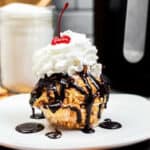 Air fryer fried ice cream on a white plate with chocolate sauce, whipped cream, and a cherry on top.
