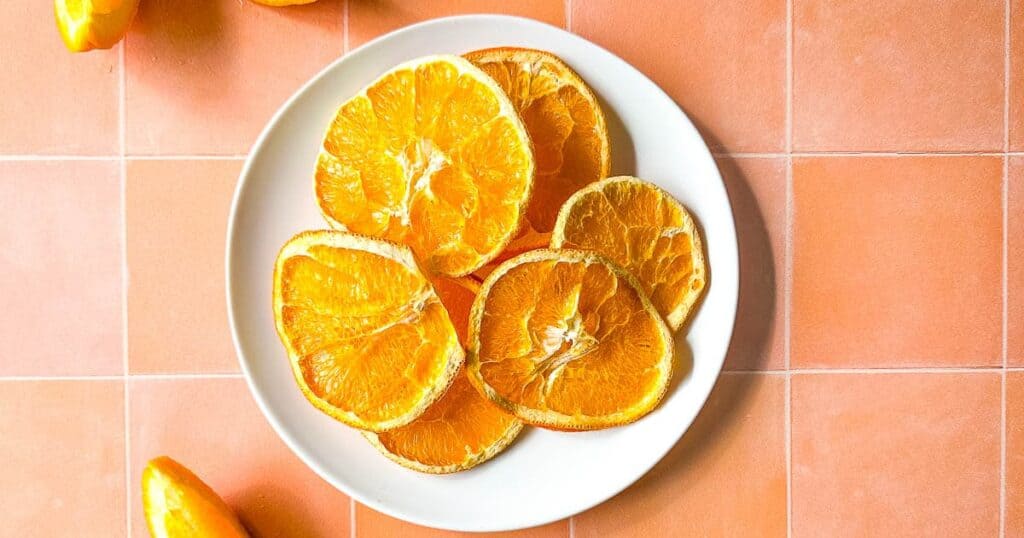 Air fryer dehydrated orange slices on a white plate surrounded by fresh orange slices.