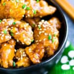 Fried chicken pieces in orange sauce garnished with sesame seeds in a black bowl with chopsticks and a green and white napkin.