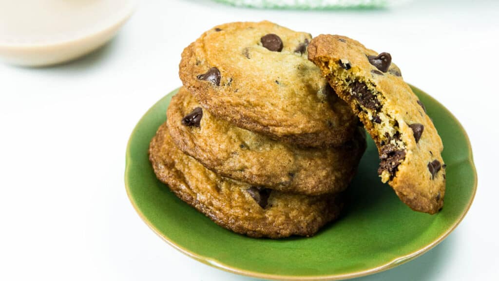 A stack of three cookies with chocolate chips sit on a green plate, while another cookie with a bite missing leans on the stack.