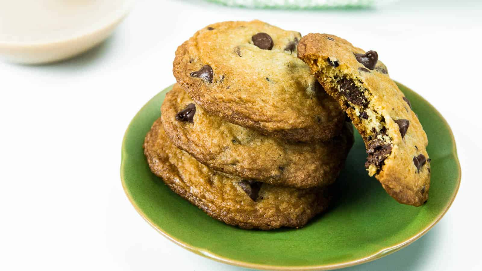 A stack of three cookies with chocolate chips sit on a green plate, while another cookie with a bite missing leans on the stack.