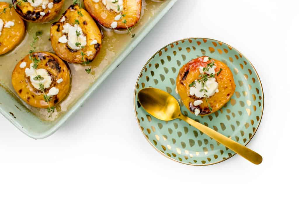 Baked peaches in a green baking dish next to a speckled plate with a baked peach on it. 