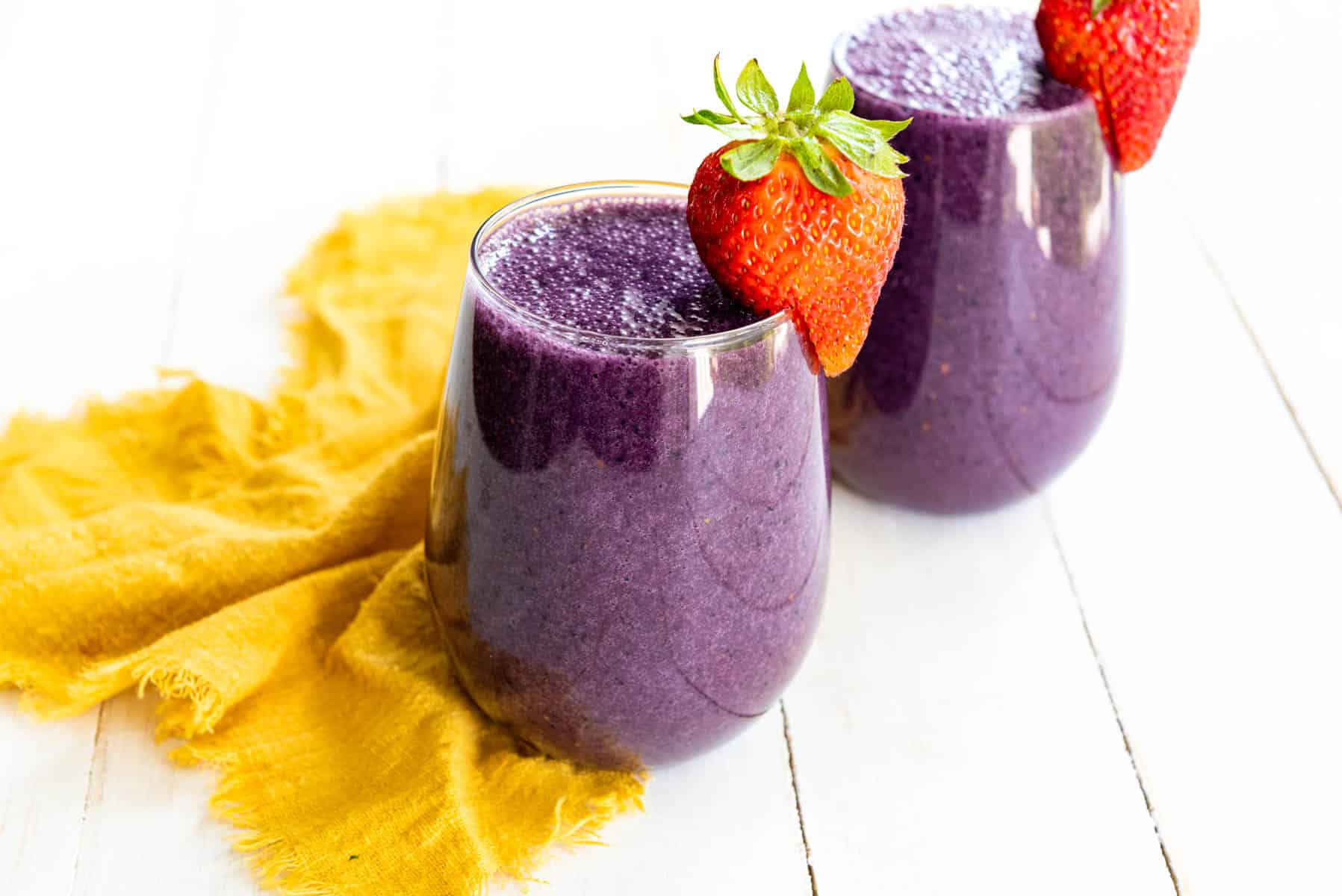 two purple smoothies in stemless wine glasses with a fresh strawberry garnish.
