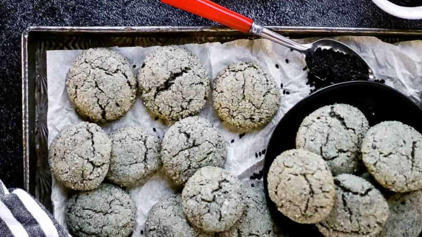 Black sesame cookies on a baking sheet with a spoonful of black sesame seeds.