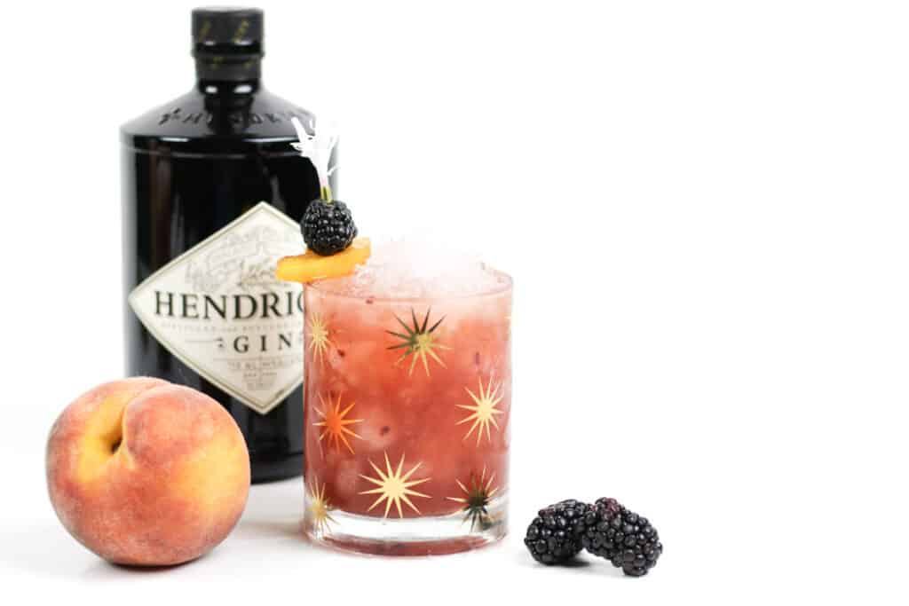 A bottle of gin next to a peach, blackberries and a peach blackberry cocktail.