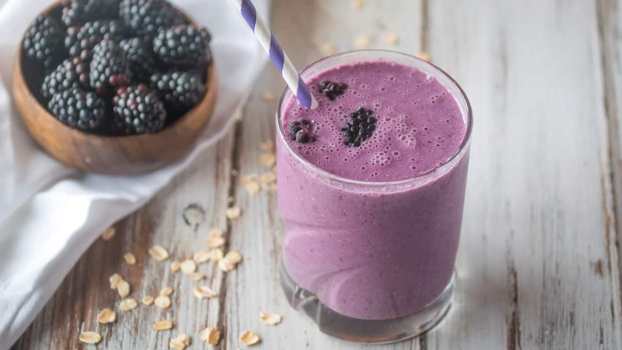 Blackberry smoothie in a glass with a bowl of blackberries.