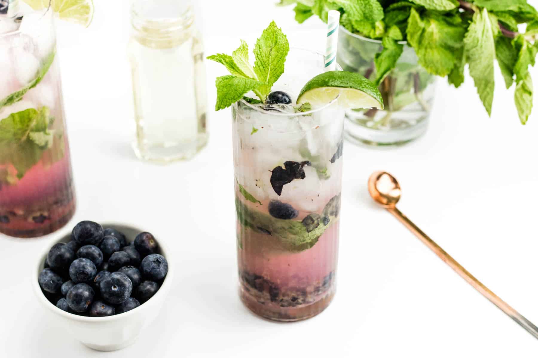 A blueberry mojito sits next to a bowl of fresh blueberries, a jar of simple syrup and a bunch of mint.