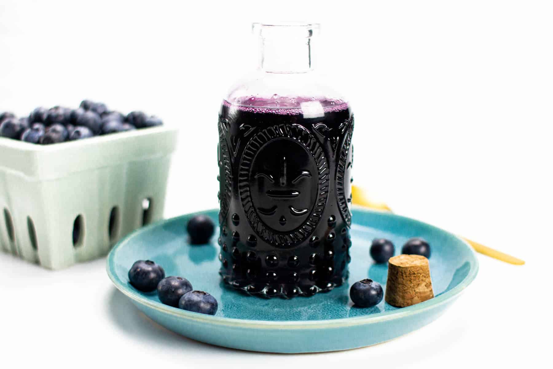a blue plate under a bottle of blueberry simple syrup next to a berry bowl of fresh berries.
