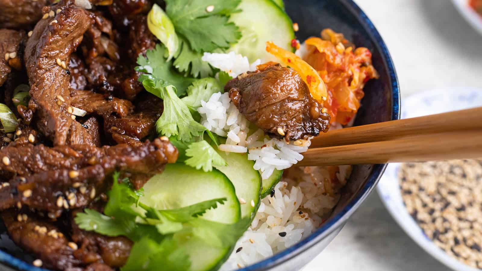 Beef bulgogi in a bowl with rice, cucumbers, cilantro and chopsticks.