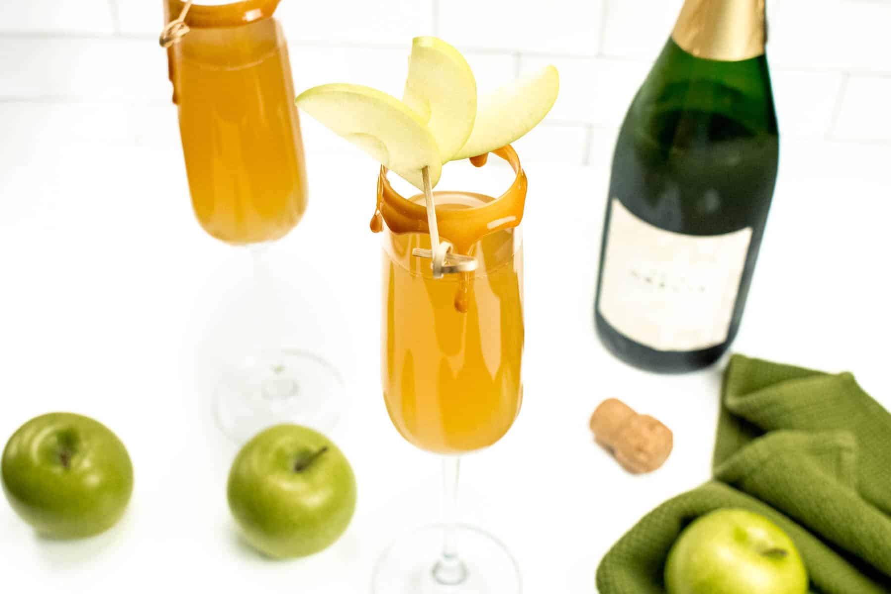 Ingredients for a caramel apple mimosa sit surrounding the finished cocktail on a white background.