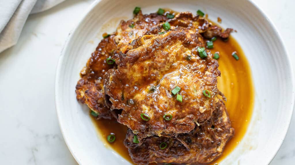 Egg foo young patties on a white plate with savory sauce and sliced green onions.