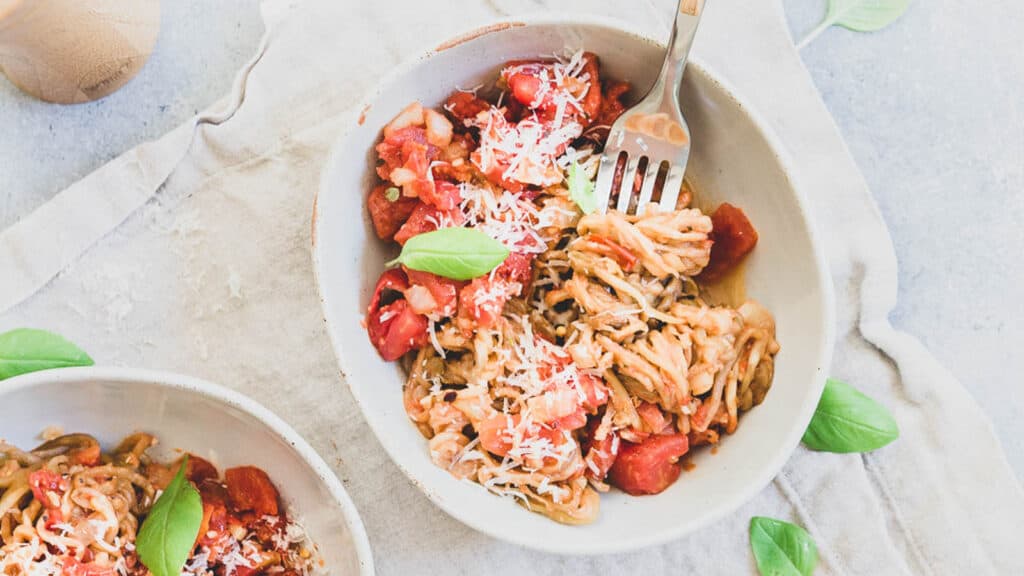 Tomato basil eggplant noodles in bowls on a neutral kitchen towel with fresh basil leaves.