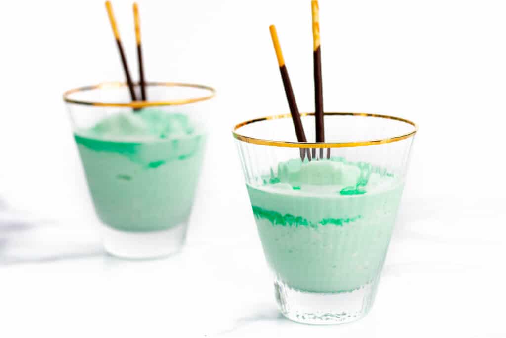 two mint green frozen ice cream cocktails with layers of liqueur and two chocolate biscuit sticks in stemless martini glasses.
