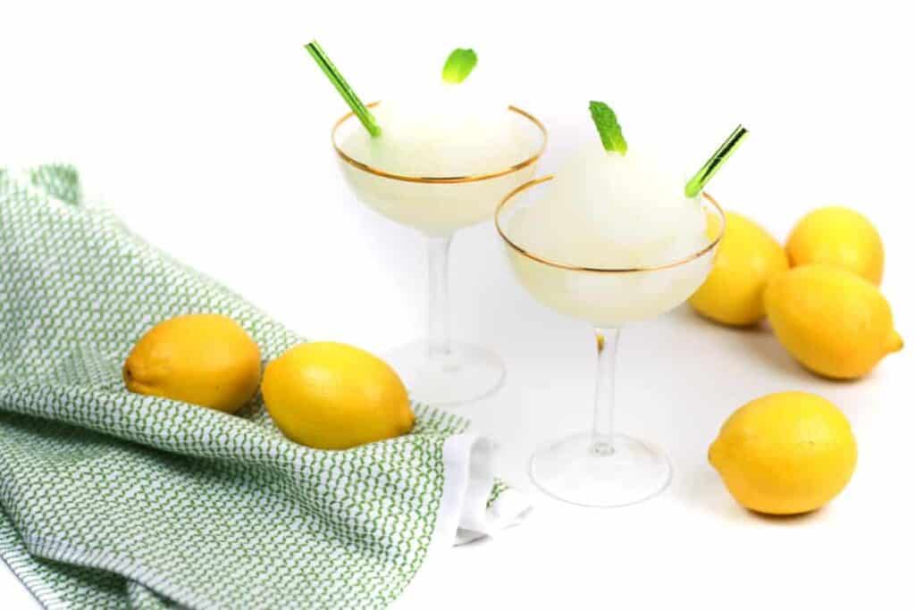 two frozen lemon cocktails in coupe glasses with gold rims, next to lemons and a green kitchen towel.
