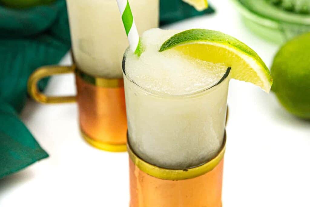 A frozen Moscow mule in a copper and glass mug with a green and white striped paper straw and a wedge of lime on a white background with a green glass citrus squeezer and a green kitchen towel.