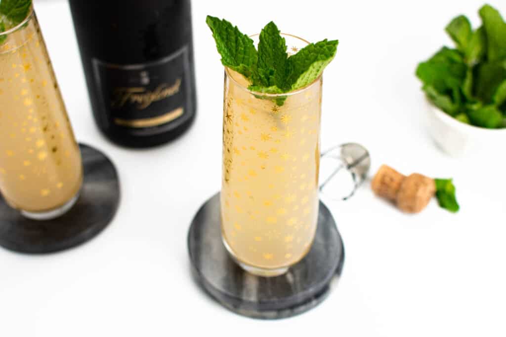 Guava mimosas garnished with mint leaves sit in stemless champagne flutes on gray coasters. A black bottle of champagne is visible in the background with the cork and cage laying alongside with additional mint leaves