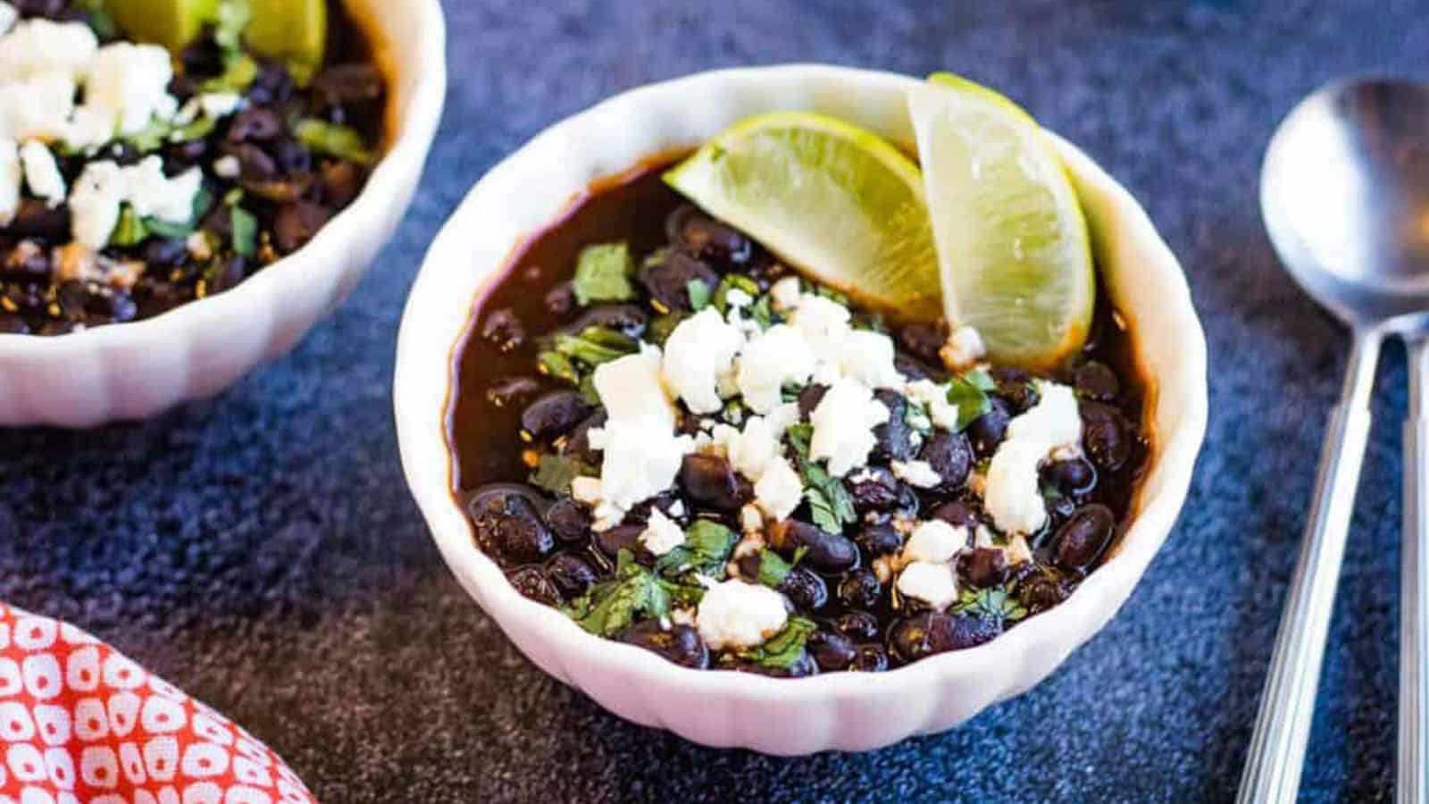 Low angle shot of two bowls of Mexican black beans garnished with crumbled cheese and lime wedges.