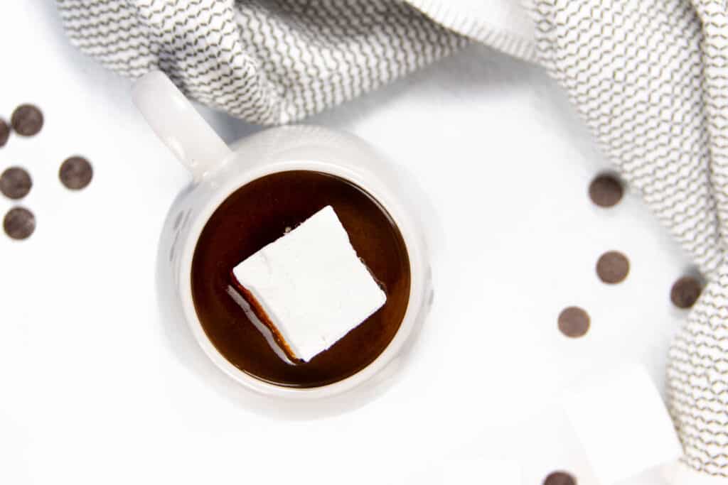 A gray mug containing instant pot hot chocolate with a gray kitchen towel, marshmallows and chocolate chips on a white background.