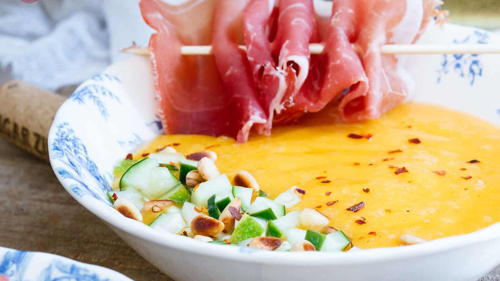 Mango melon soup with pickled cucumber and prosciutto skewer in a bowl.
