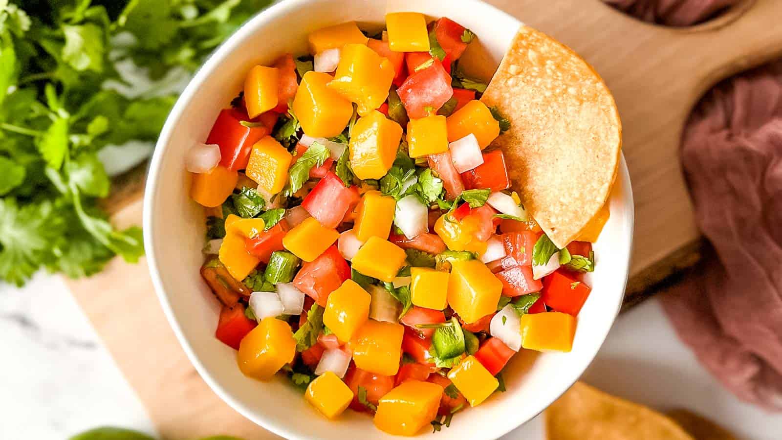 A bowl of mango pico de gallo sits on a rustic wooden cutting board surrounded by limes, cilantro, and a pink linen.