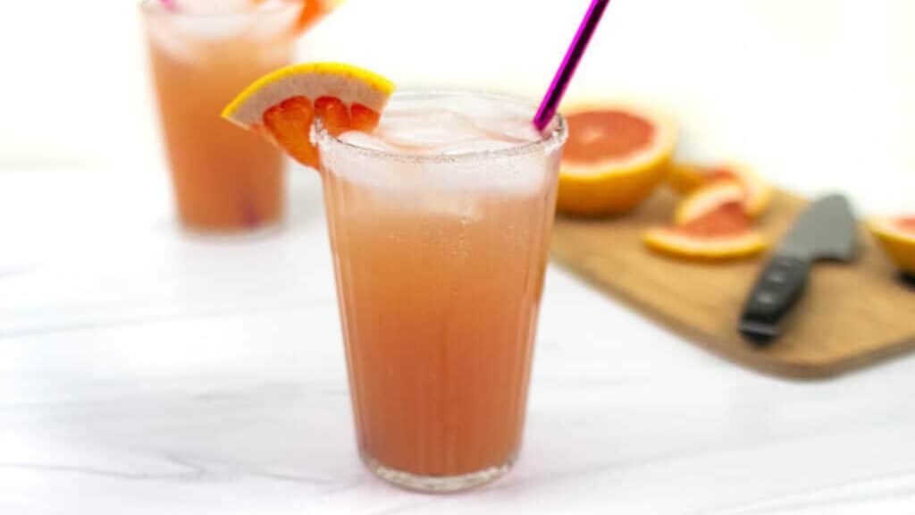 Two pink cocktails garnished with a wedge of grapefruit sit alongside a cut grapefruit on a cutting board with a knife.