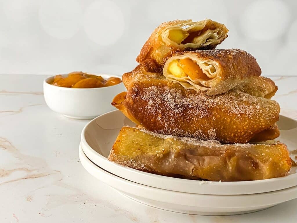 Peach cobble egg rolls set on a stack of two white plates. Peach cobbler stuffing in a separate bowl sits next to this plate.