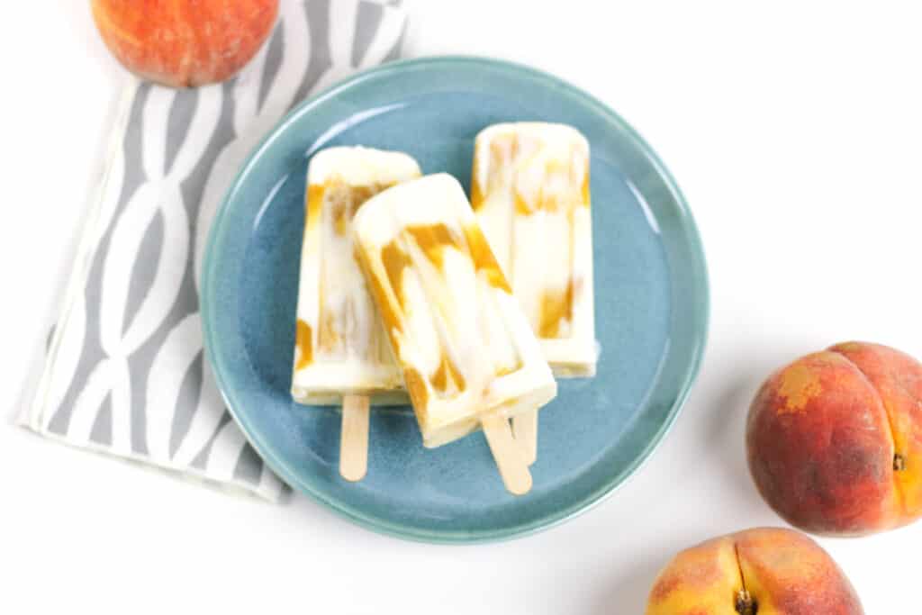 Peaches and cream popsicles on a blue plate sit with whole peaches surrounding them on a white background.