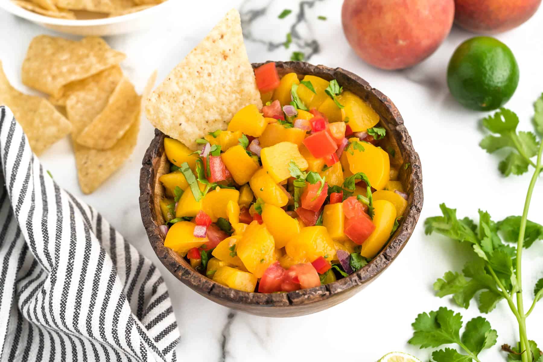 A bowl of peach salsa with a chip in the bowl, surrounded by cilantro, peaches and chips.