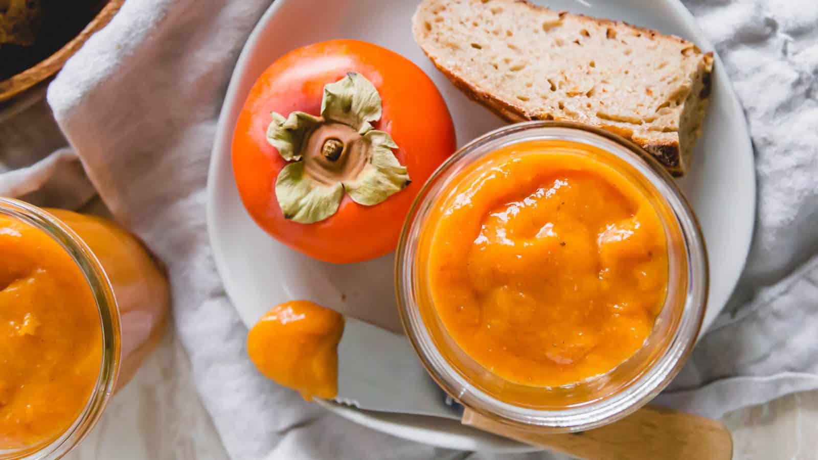 Persimmon jam in a glass jar with persimmons and toast on the side.