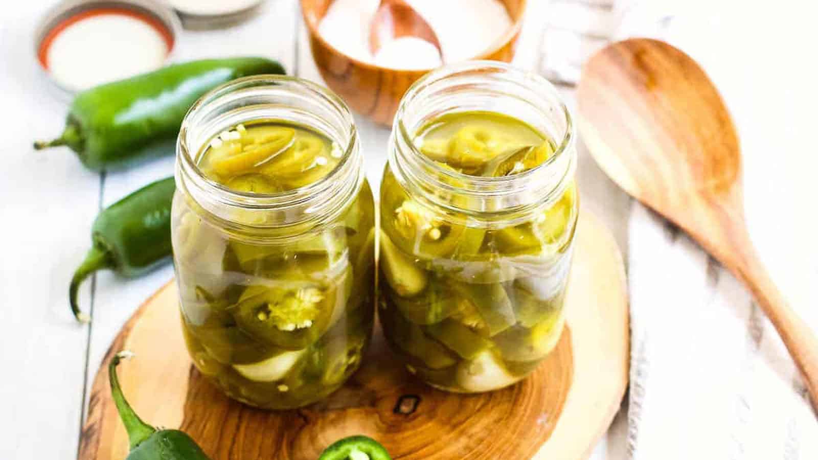 Two canning jars filled with pickled jalapenos on a wooden board with fresh jalapenos on the side.